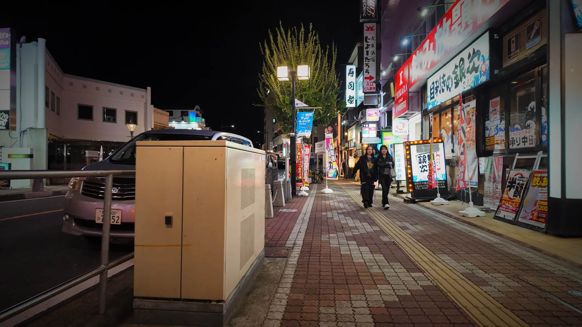 Two people chatting as they walk along the lively station-front street at night, with the glow of izakayas behind them
