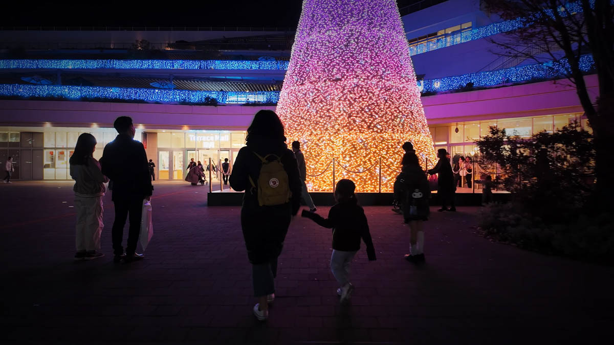 A giant Christmas tree in the station plaza at night, with passersby stopping to take photos