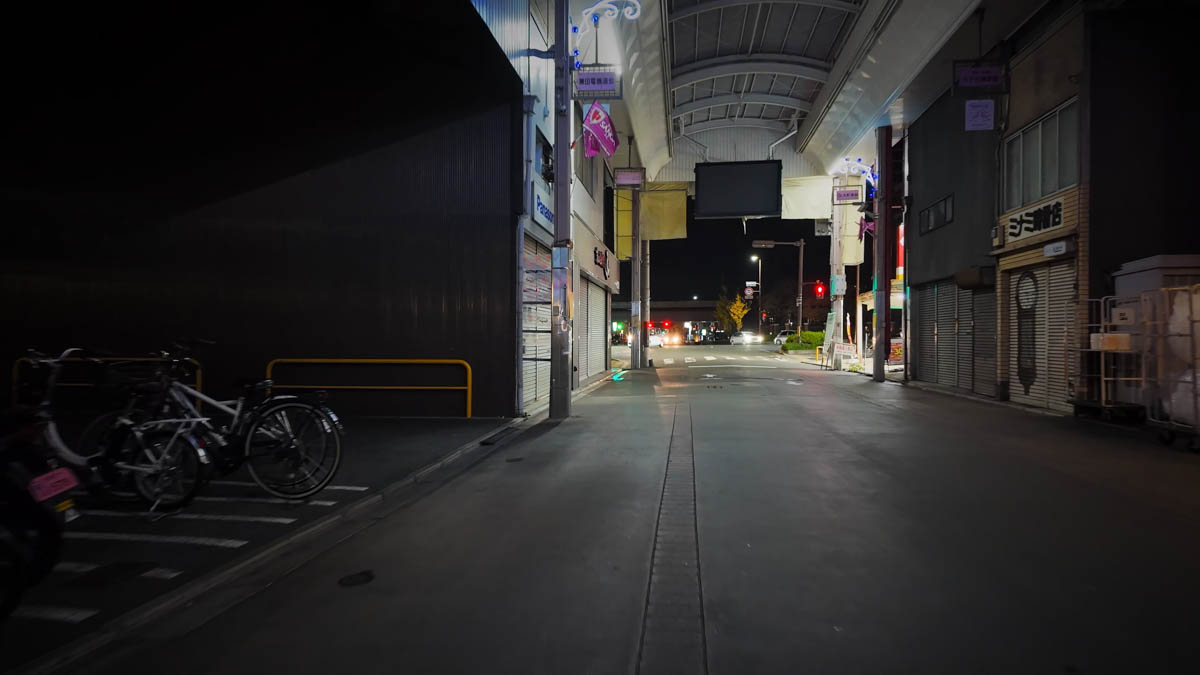 A night street leading back toward the main road, with bicycles and shop signs along the way