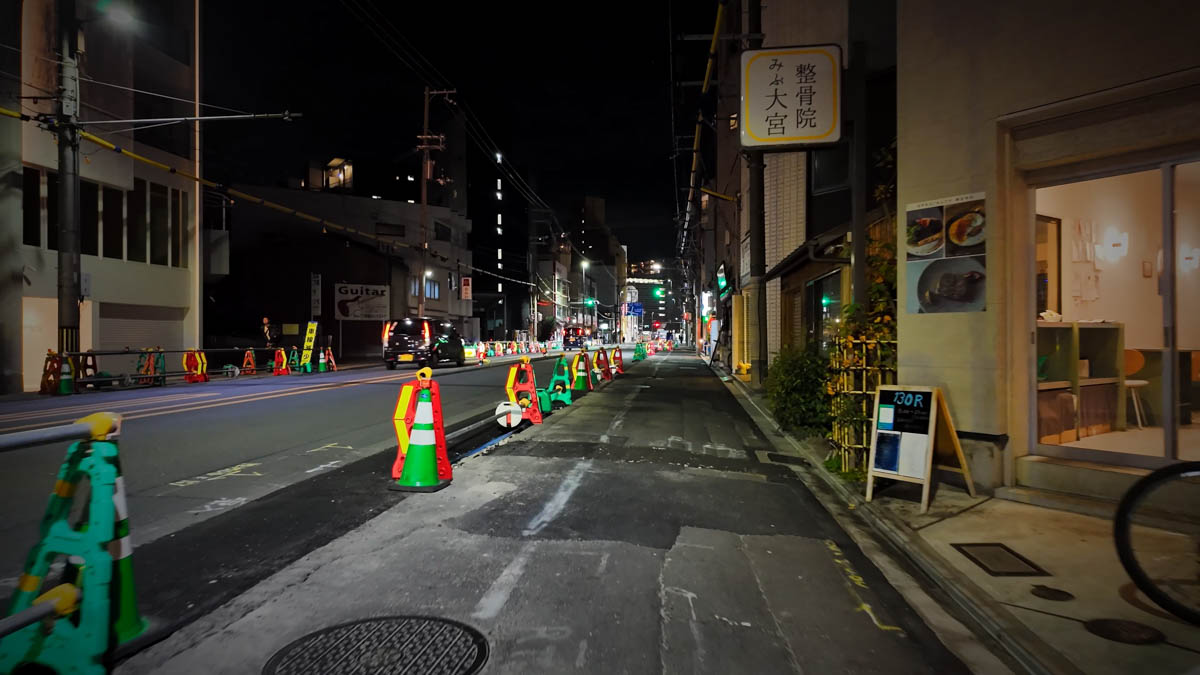 A street under construction at night, with traffic cones and a small lit shop along the sidewalk