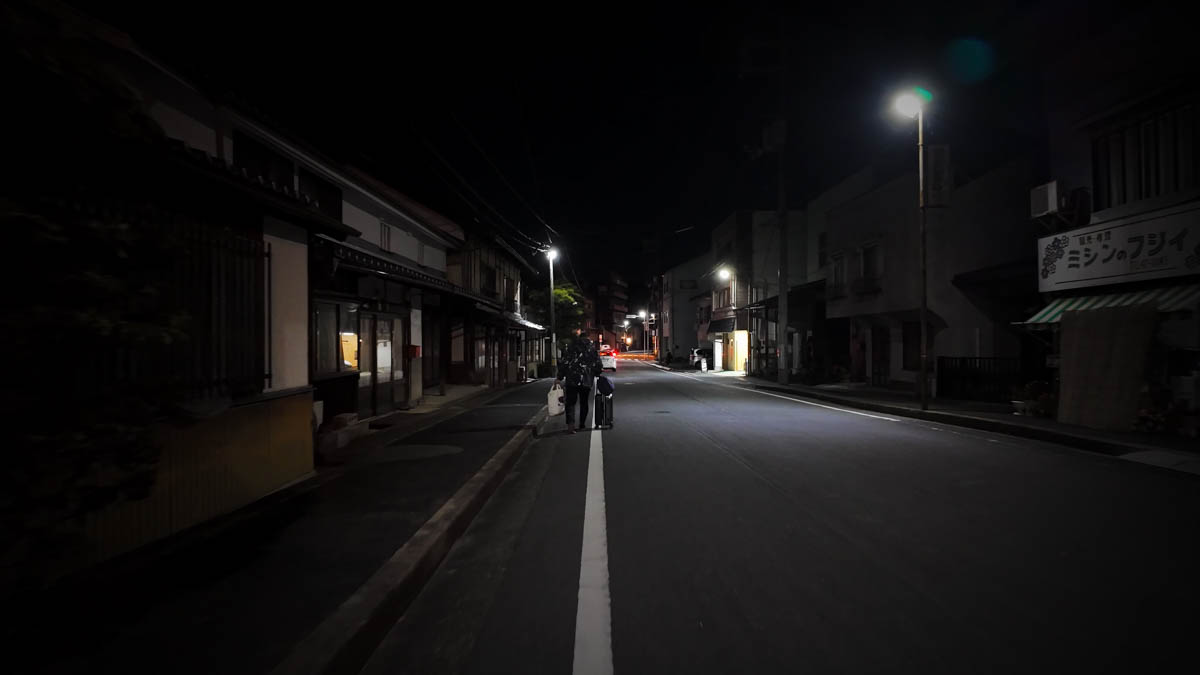 A night street near Kuse Station, with old buildings, a Fujii Sewing Machines sign, and one person standing by the road