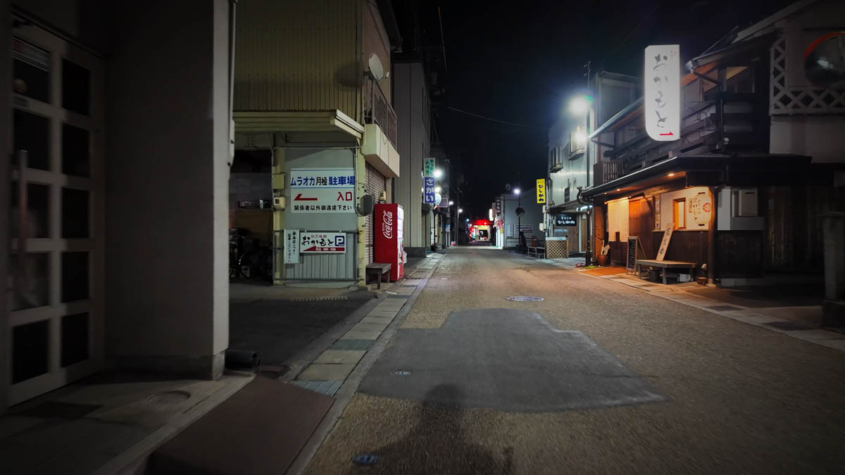 A street outside the arcade, with the Okamoto sign, a Coca-Cola vending machine, and a monthly parking sign
