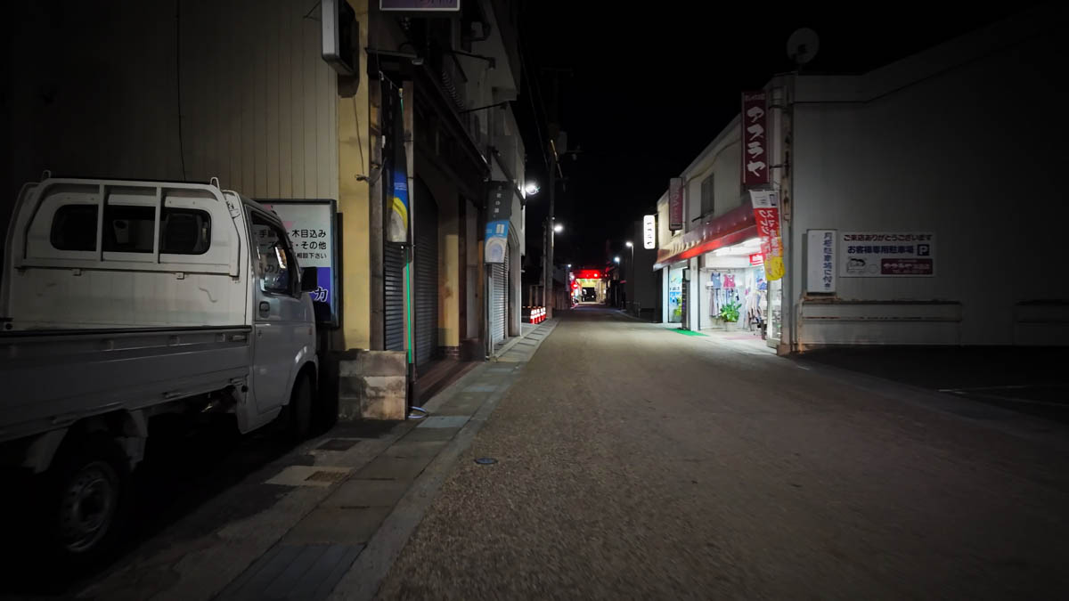 A side street with a small truck parked on the left and a lit shop on the right
