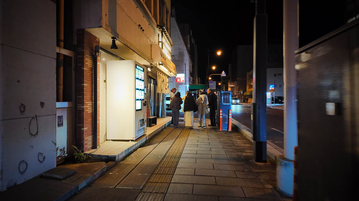 People gathered on the sidewalk along Imazuyabashi-dori at night, in front of restaurants and small shops