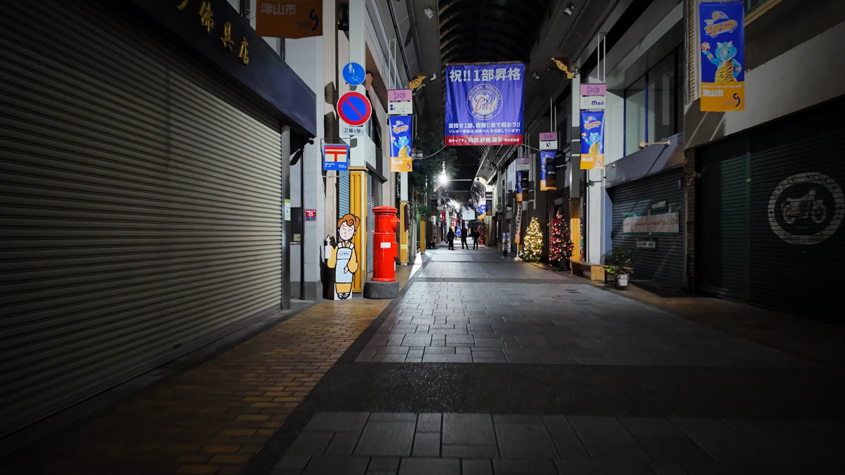 A covered shopping arcade in Tsuyama, with closed shutters, a red cylindrical post box, and small illuminations