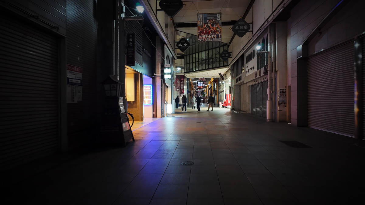 A covered shopping arcade at night, with a lit shop and a few pedestrians visible