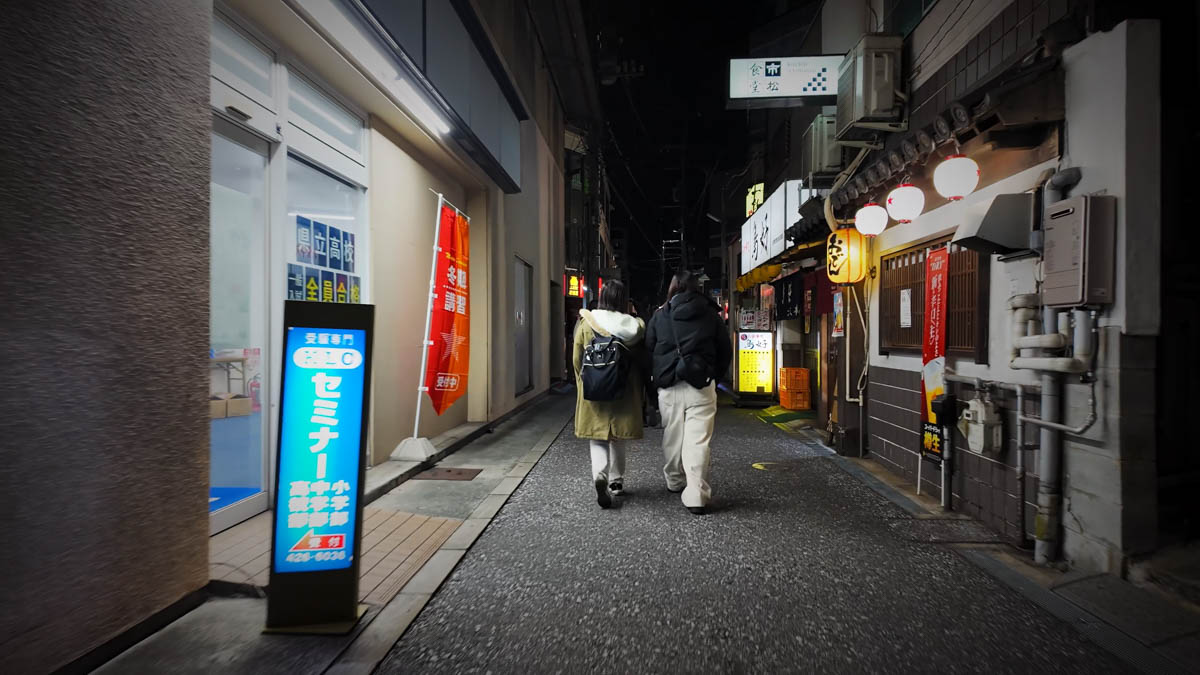 A narrow night street with cram school signs, izakaya lanterns, and two people walking ahead