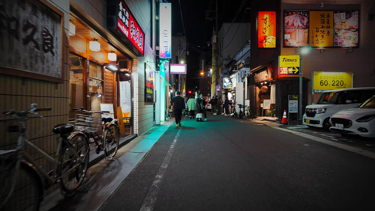 A night street in Kurashiki's entertainment area, with restaurant signs, a full parking lot, and several pedestrians