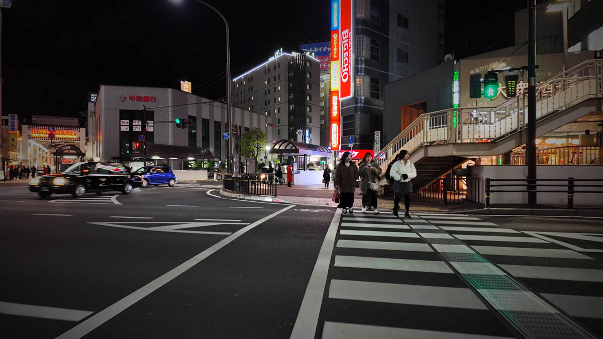 A night intersection in front of Kurashiki Station, with the Chugoku Bank sign, BigEcho lights, and people crossing the street