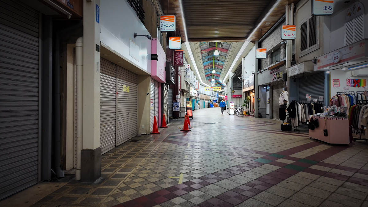 Inside a covered arcade in Fuse at night, with shuttered shops alongside a clothing store and the passageway continuing ahead