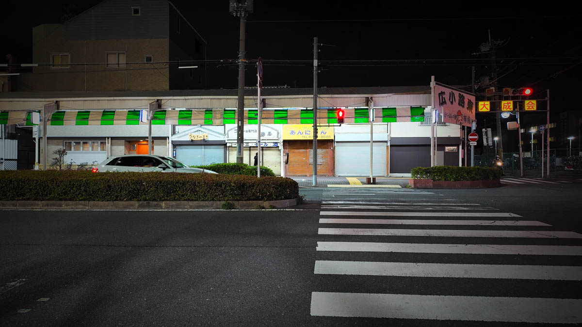 Near the entrance to Hiroko-ji Shopping Street, with the arcade sign and shuttered shops visible beyond a crosswalk
