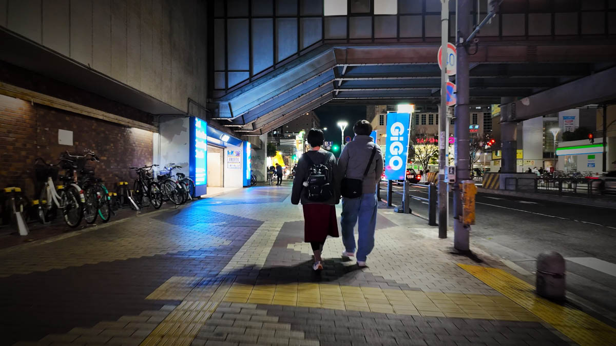 Near the elevated tracks at Fuse Station, with pedestrians on the sidewalk, a bicycle parking area, and streetlights along the road