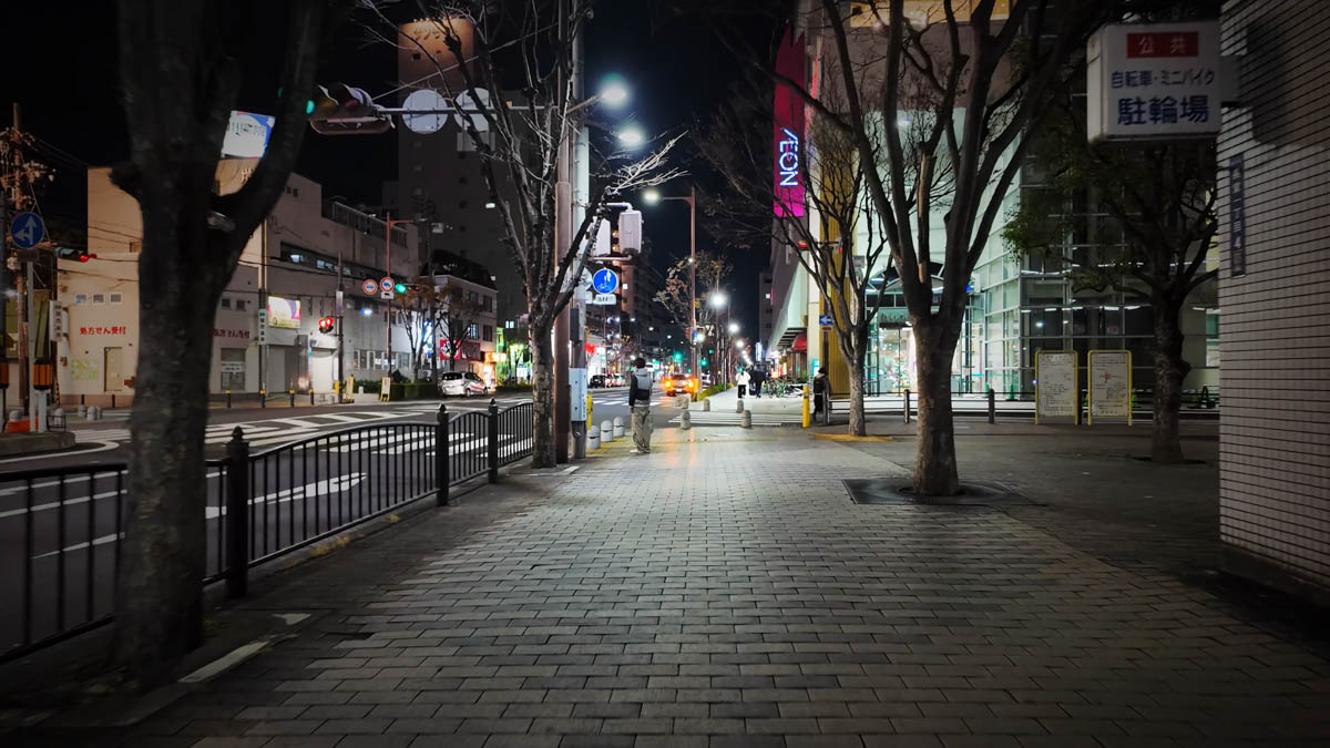A wide sidewalk in front of Fuse Station at night, lined with street trees, the Aeon building, and the road ahead