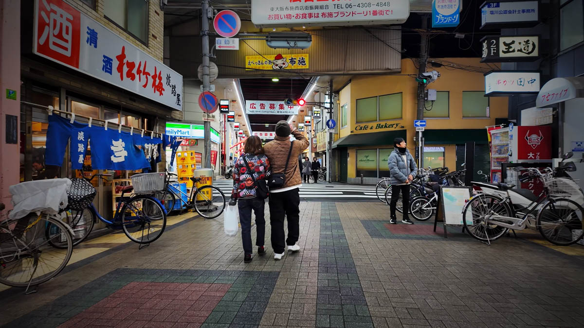 Near the entrance to a shopping arcade in Fuse, with an izakaya curtain, a FamilyMart, and the arcade arch