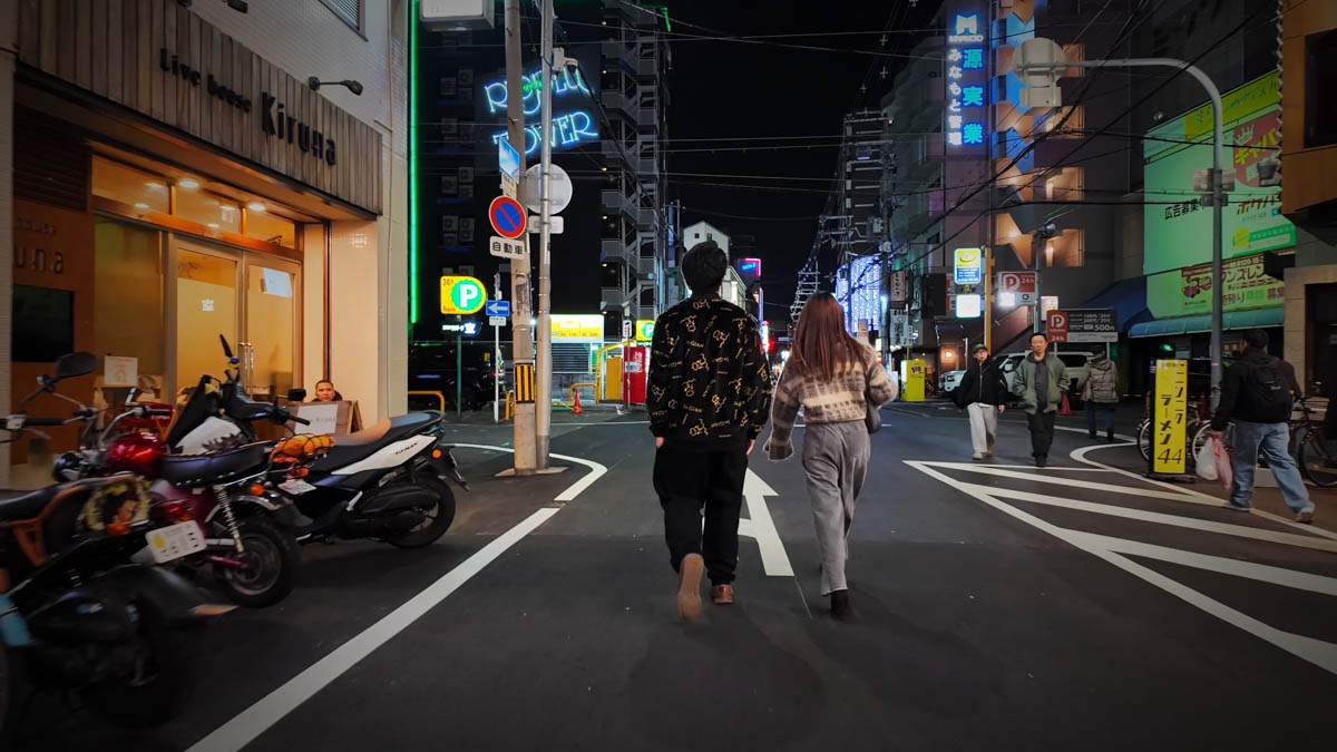 A night street in Fuse, with restaurant signs and lights on either side, and a couple walking ahead