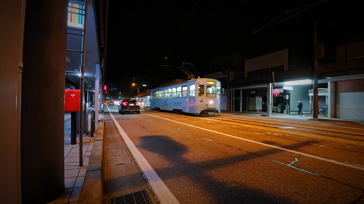 A Manyo Line tram running along a night street. The light blue car has its headlights on. A red postbox stands to the left