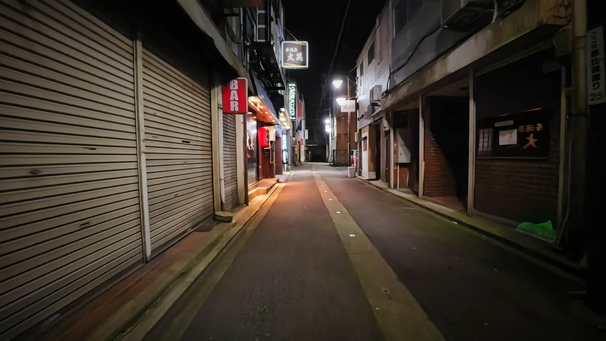 A narrow alley with shuttered storefronts and signs for a bar and a charcoal-grill restaurant still visible