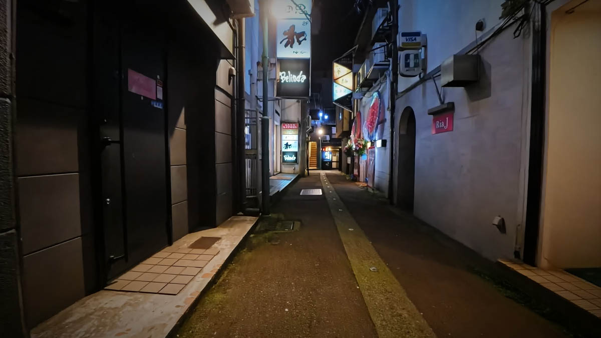 A narrow alley lined with snack bars and clubs. Signs for Belinda, Wa, and Ria J are visible