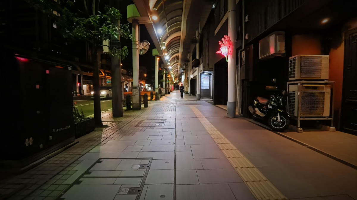 The end of a covered arcade, with a red neon decoration and a parked bicycle. A lone figure walks in the distance