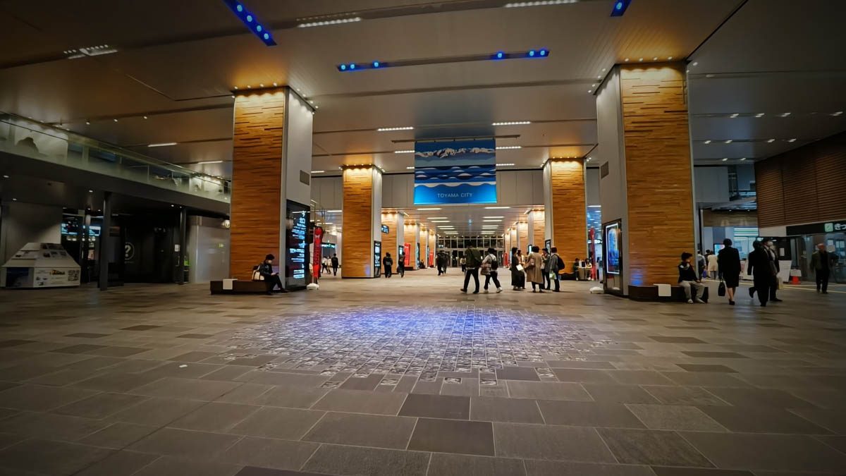 富山駅構内へ続く広い通路。木目調の柱と青い天井照明、富山市の案内板が見える