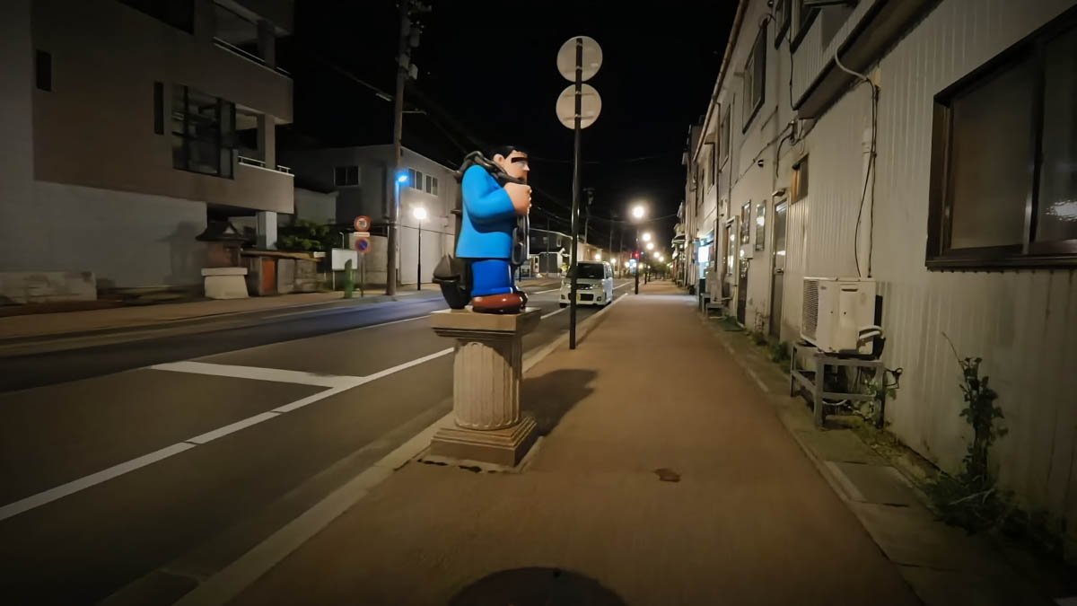 夜の通り。台座の上に怪物くんのお供フランケンの像が立ち、左右に住宅と店舗が並ぶ