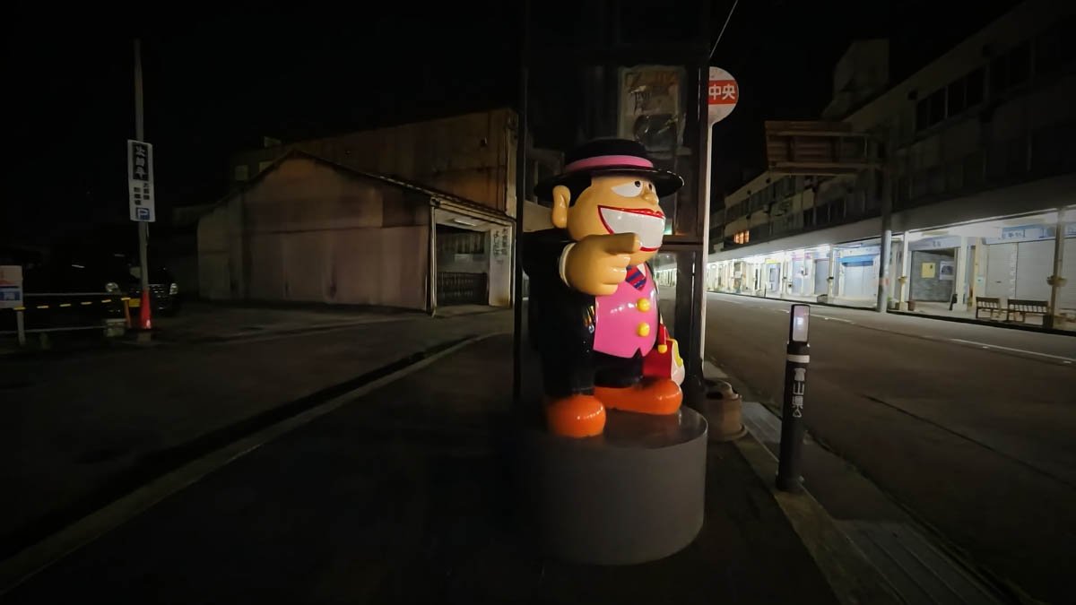 夜の氷見駅前バス停。「中央」の停留所標識の横に、笑ゥせぇるすまんの喪黒福造の像が立つ