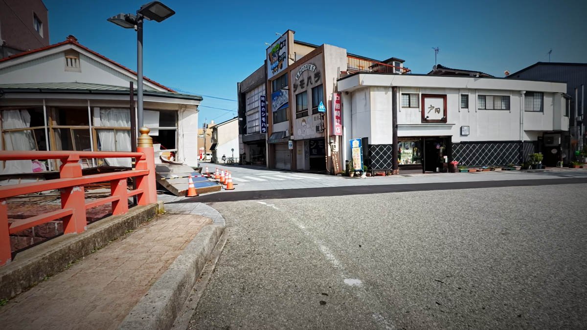 七尾市内 橋と交差点 地震被害 赤い欄干の橋の近く。左の建物が崩れ、シモハラの看板が残る交差点。路面には土砂とコーンが置かれている