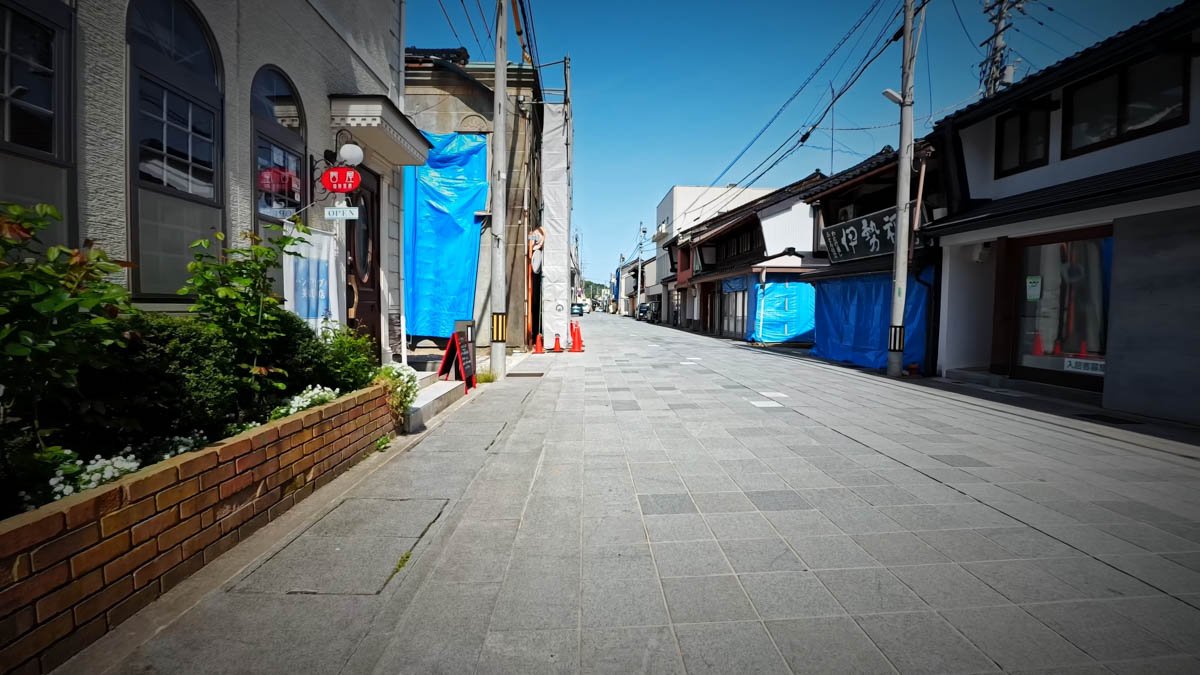 七尾 一本杉通り 一本杉通り。左手の洋風の建物にOPENの看板と花壇、外壁にはブルーシートがかかる。右手の町家建築にもブルーシートがかかり、奥に続く通りにコーンが見える