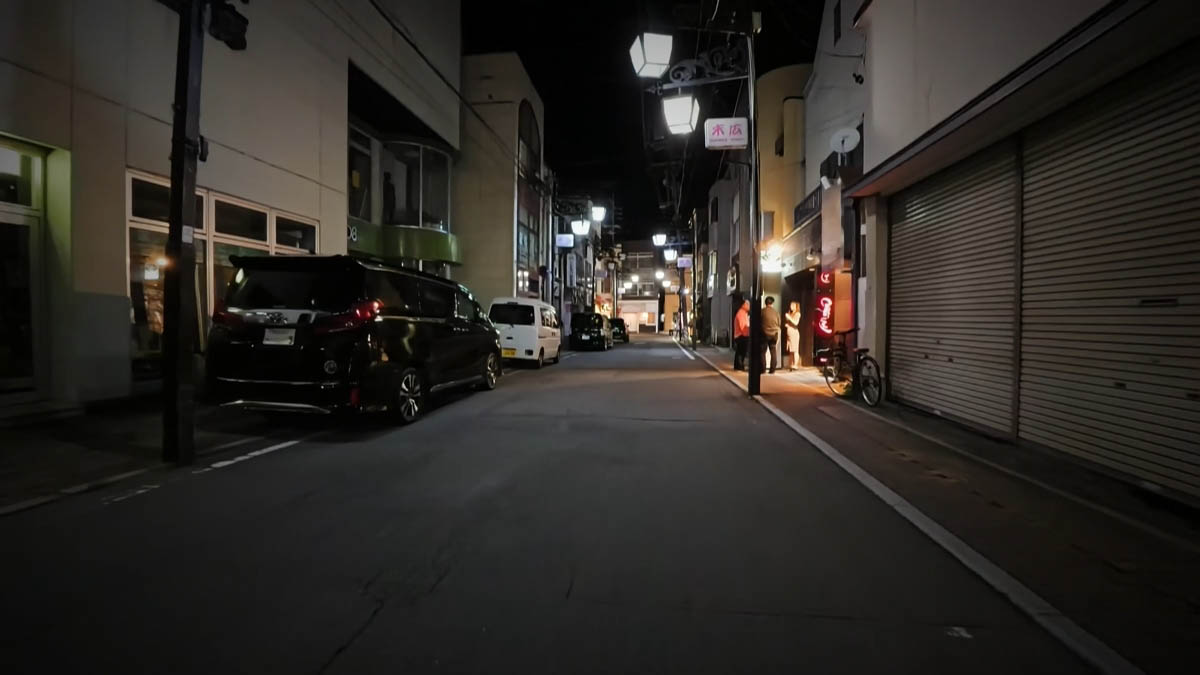 末広付近 シャッターの店と自転車。奥に明かりの店先で立ち話する人、末広の案内