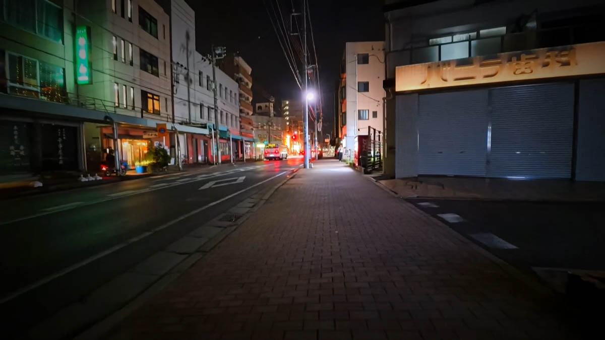 Station-front street Brick sidewalk and dental clinic sign; a bus and streetlamp ahead, hotel and chuka soba signs across the street
