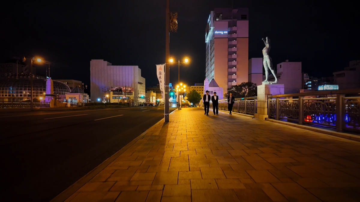 People walking on Nusamai Bridge toward a Four Seasons statue and Dormy Inn