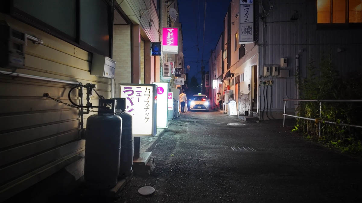 ガスボンベの脇に立つパブ・スナックの看板と、ヘッドライトのタクシー